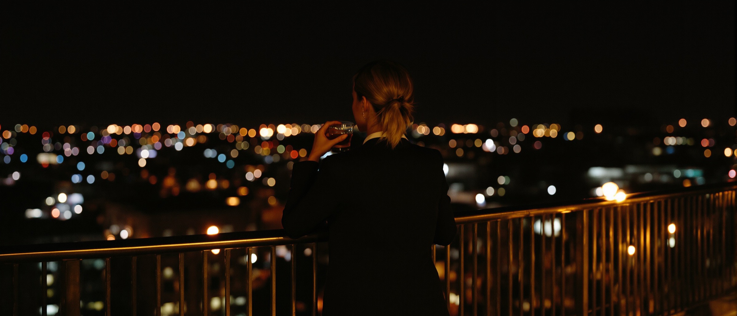 A woman in a dark blazer leaning on a rooftop railing at night, city lights out of focus behind her.