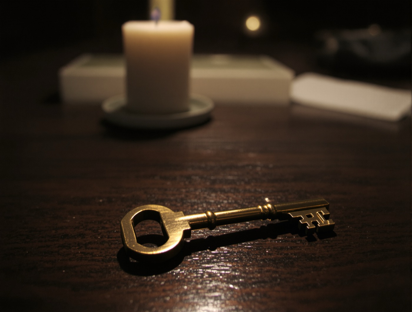 A brass hotel key on a dark walnut reception desk beside an unlit candle, under one warm spotlight.