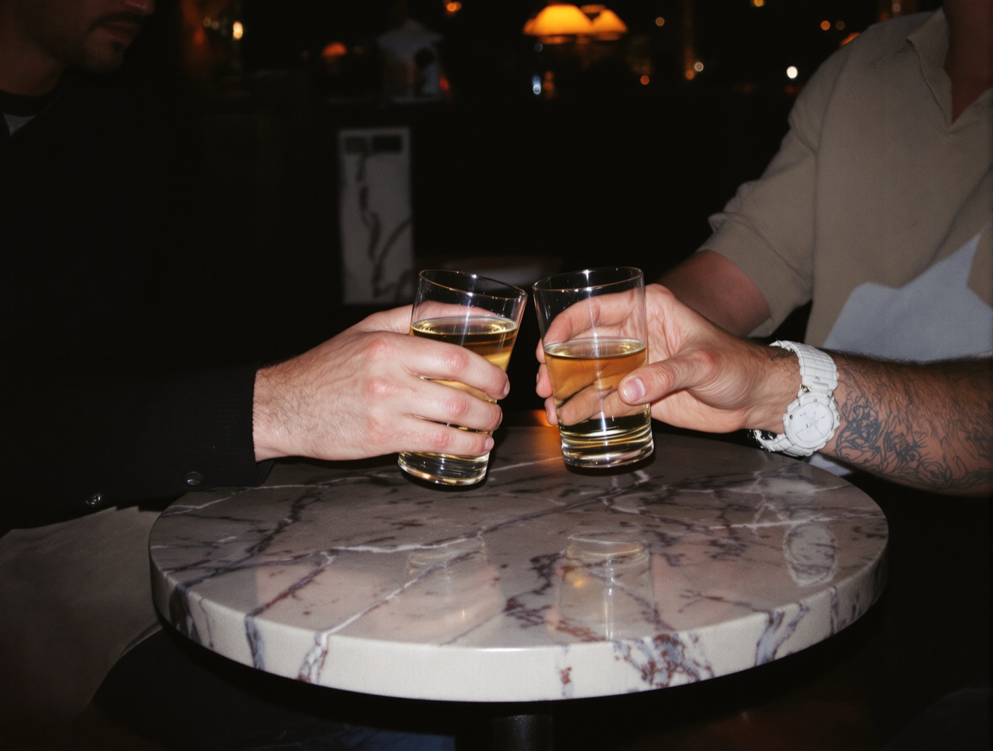 Two people touching short glasses together over a small marble table in a dim lounge.